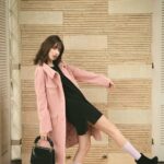 Fashion model in pink coat and black dress striking a dynamic pose indoors with stylish black heels.